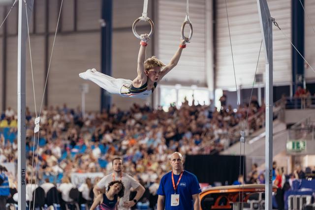 Young male gymnast holds a cross position on still rings, intense focus, crowd-filled arena in background.