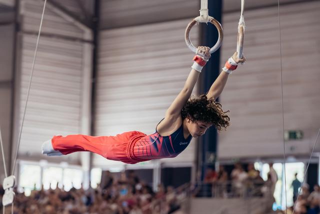 Young male gymnast performs an iron cross on rings, body horizontal, intense focus, in a packed indoor arena.
