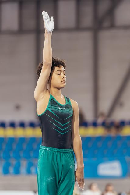 Young male gymnast stands with eyes closed, one arm raised in salute, wearing teal and black leotard in arena.