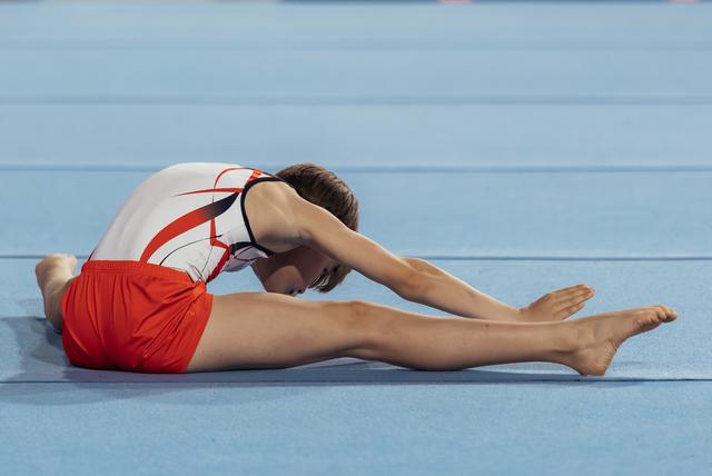Young male gymnast in red and white leotard performs a seated forward stretch on a blue competition floor, head bowed.