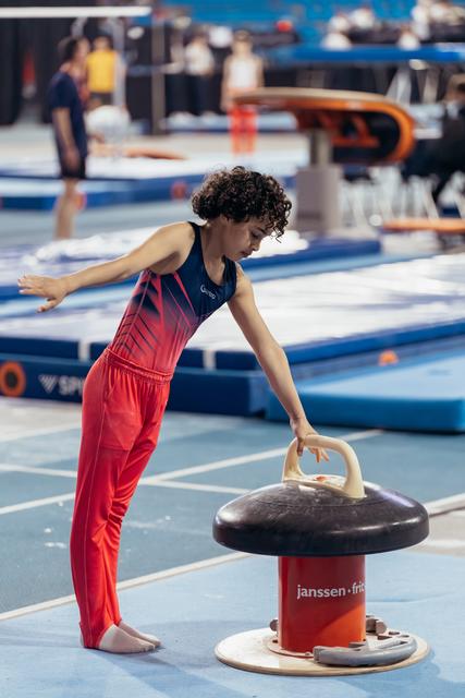 Young female gymnast with curly hair leans over pommel horse, focused and deliberate, in a busy competition hall.
