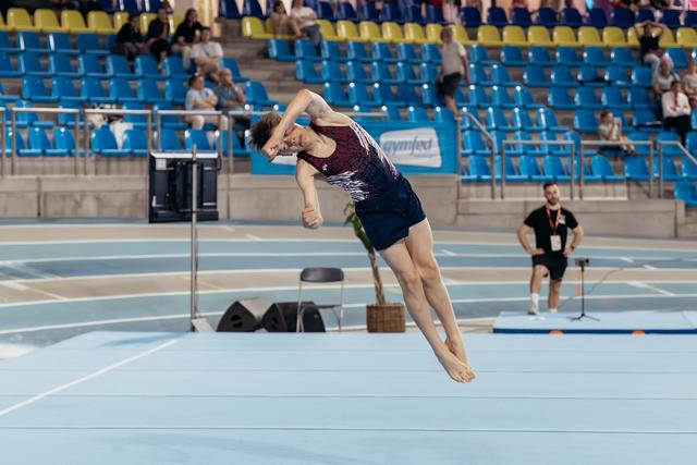 Male gymnast mid-air during floor exercise, body tucked tight, intense focus, indoor arena with blue floor