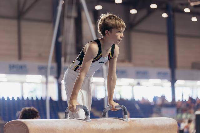 Young gymnast stands focused on pommel horse, gripping handles with determined expression in a sports hall.