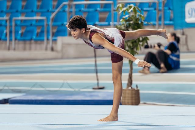 Young male gymnast holds a precise arabesque balance on the floor, focused expression, in a blue-seated arena.