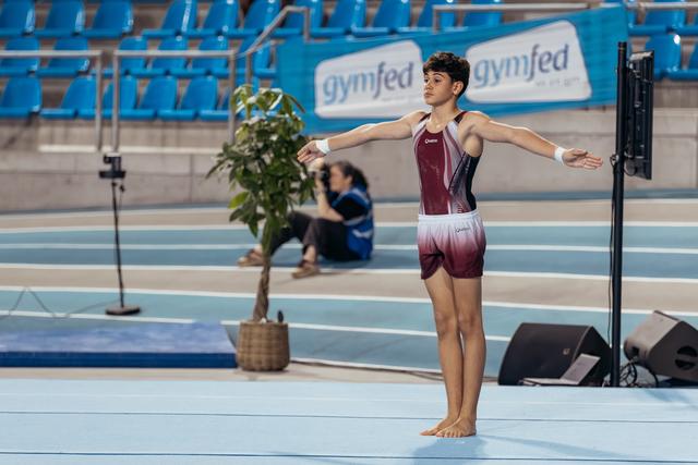 Young male gymnast stands barefoot on floor mat, arms wide open, composed expression, during a Gymfed indoor event.