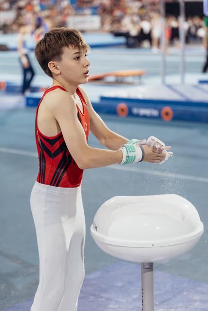 Young male gymnast applies chalk to his hands with focused concentration, wearing red and black leotard at a competition arena.