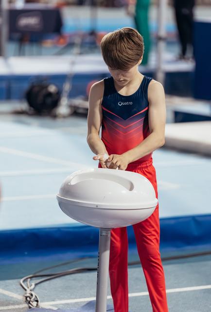 Young male gymnast in red and navy leotard applies chalk to hands at a chalk bowl, focused before his routine.