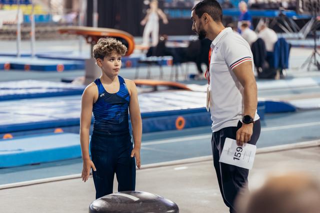 Young male gymnast listens attentively to coach's instructions before vault, both standing on competition floor.
