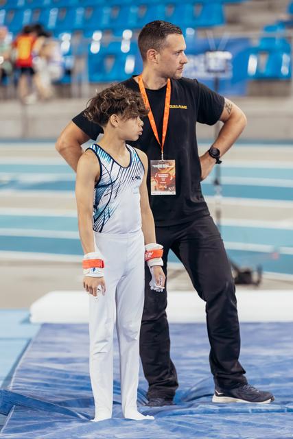 Young gymnast stands focused beside coach in black, both observing the floor exercise area at an indoor sports arena.