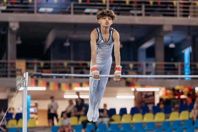 Young male gymnast grips the high bar with focused intensity, body extended, wearing red wrist guards in a sports arena.