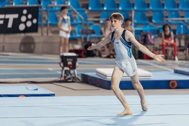 Young male gymnast performs floor routine, arms extended with focused concentration in a blue arena setting.