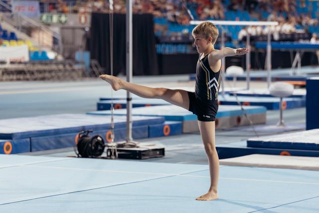 Young male gymnast performs a precise leg raise on the floor exercise mat, arms extended, showing strong balance and focus.