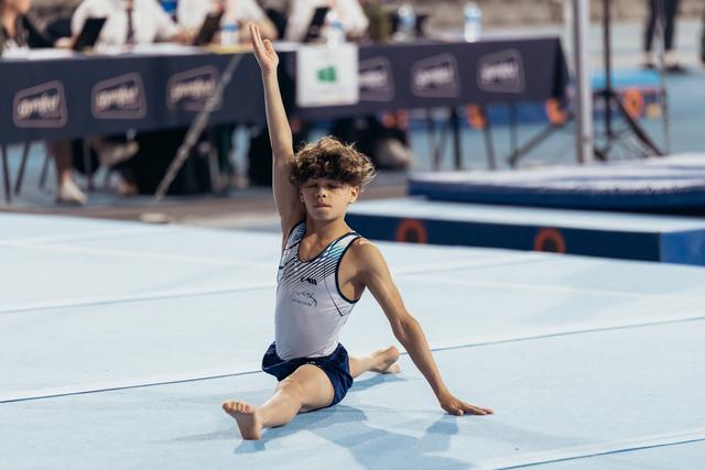 Young male gymnast holds a graceful seated pose on the floor mat, one arm raised high, focused expression.
