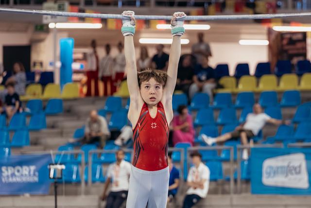 Young male gymnast hangs from the high bar with intense focus, wearing a red leotard in a packed sports hall.