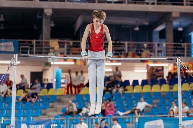 Young male gymnast grips the high bar with intense focus, body suspended mid-routine in a packed sports arena.