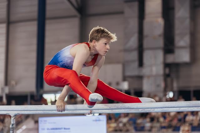 Young male gymnast performs a straddle hold on parallel bars, his focused expression intense during a competition.