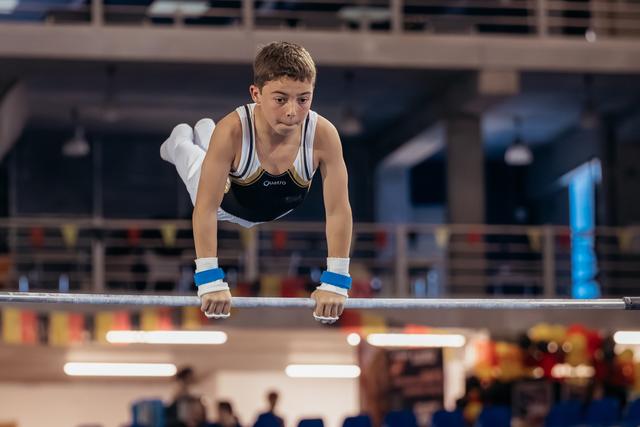 Focused teenage gymnast performs a support hold on parallel bars, wearing blue wristbands, with intense concentration.