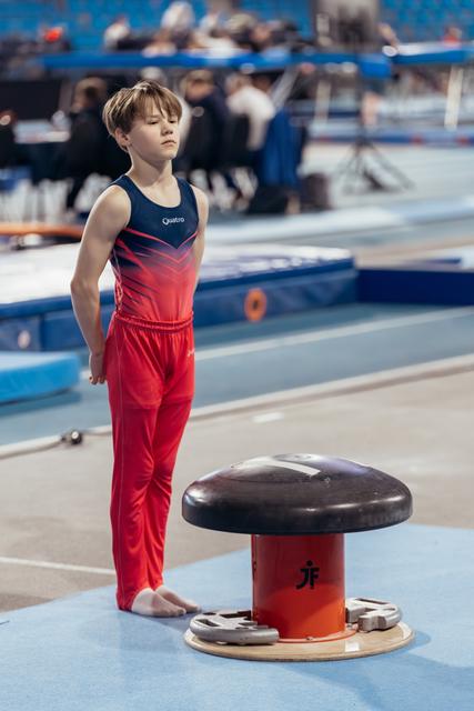 Young male gymnast in red and navy Quatro suit stands barefoot, eyes closed, composing himself before pommel horse routine.