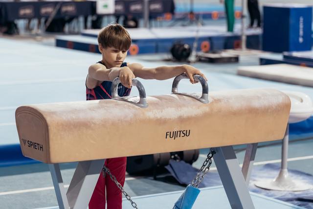 Young boy in gymnastics leotard grips pommel horse handles with intense focus in a blue competition gym.