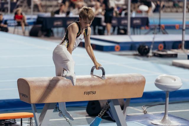 Young male gymnast leans intently over a Fujitsu pommel horse, gripping the handles with focused concentration at an indoor sports event.