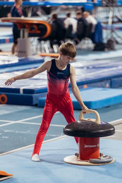 Focused young male gymnast in red uniform grips pommel horse handles, preparing for routine in blue gymnasium.