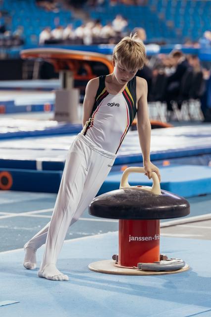 Young male gymnast in white leotard grips pommel horse handles, focused expression, blue gymnasium floor.
