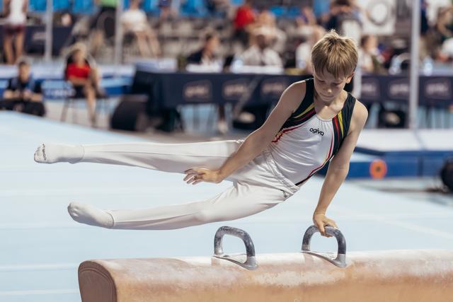 Young male gymnast performs a focused pommel horse routine, legs extended, intense concentration on his face at a gymnastics meet.