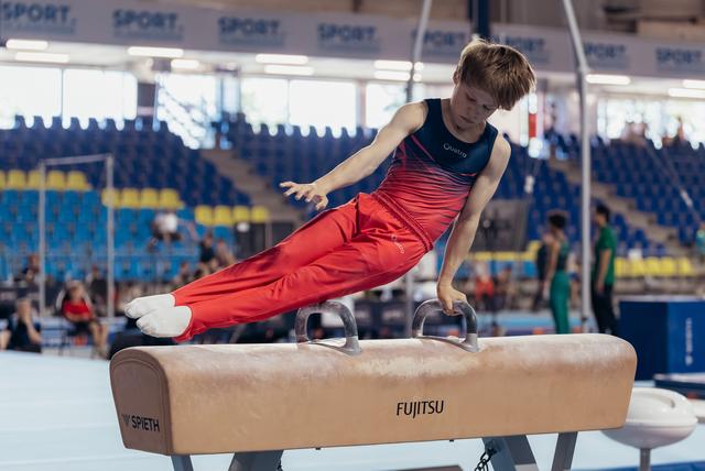 Young male gymnast performs a focused pommel horse routine in a red and navy leotard at an indoor sports arena.