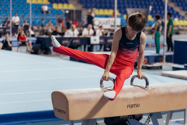 Young male gymnast performs focused pommel horse routine, legs extended, wearing red and navy at an indoor gymnastics meet.