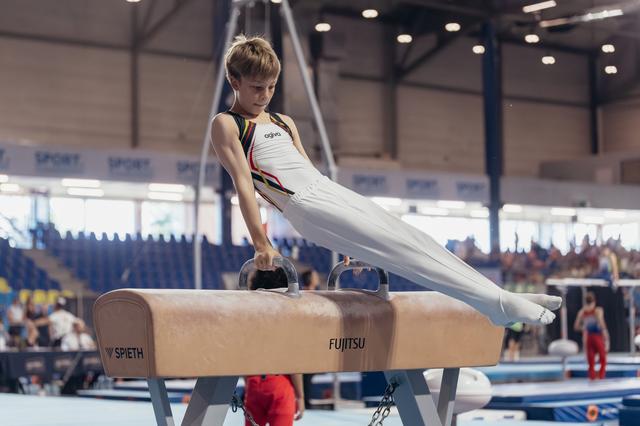 Young male gymnast performs a focused pommel horse routine in a competitive gymnastics hall, wearing a white and black leotard.