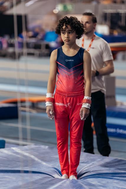 Young gymnast in red and navy Quatro leotard stands still on mat, expression serious and focused before performance.