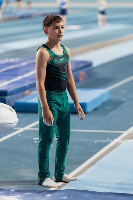 Young male gymnast stands barefoot on the floor exercise mat, looking up with focused concentration, wearing green and black attire.