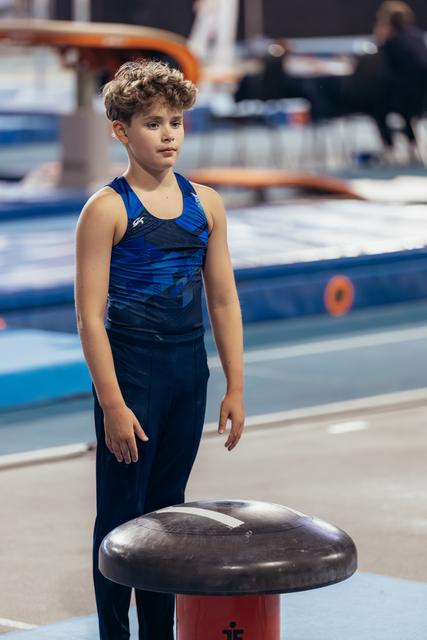 Young male gymnast stands focused beside the vault, wearing blue GK leotard, ready to compete in a gymnasium.