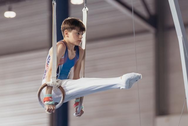 Young male gymnast performs an L-sit on still rings, wearing a blue leotard, with intense concentration on his face.