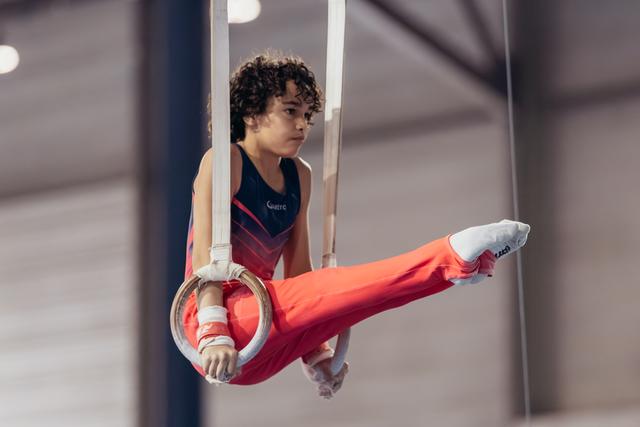 Young male gymnast holds still rings with intense focus, one leg raised, wearing red pants and navy leotard.