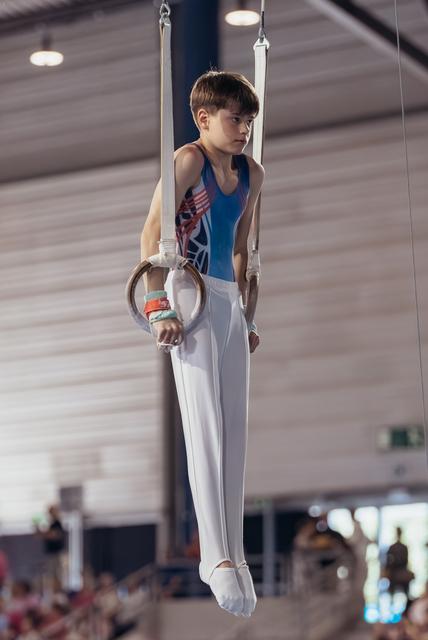 Focused young male gymnast holding still rings in iron cross position, wearing blue leotard and white pants in gymnasium.