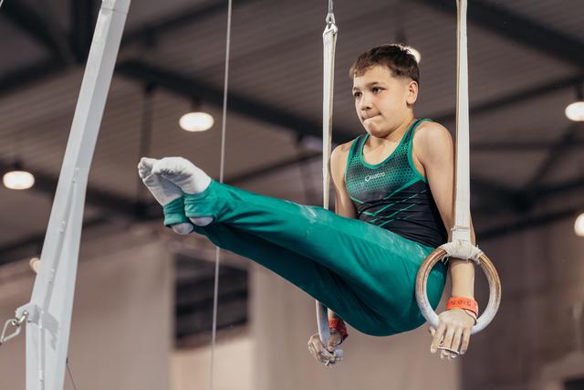 Young male gymnast holds still rings with concentration, legs raised mid-routine in an indoor gymnastics hall.