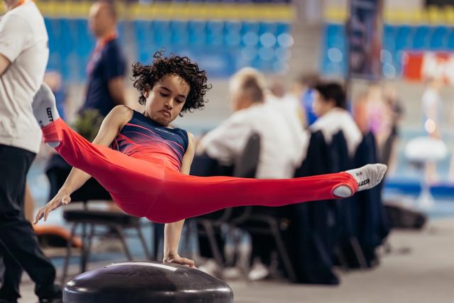 Young gymnast with curly hair performs a focused straddle hold on pommel horse, legs fully extended, intense concentration.