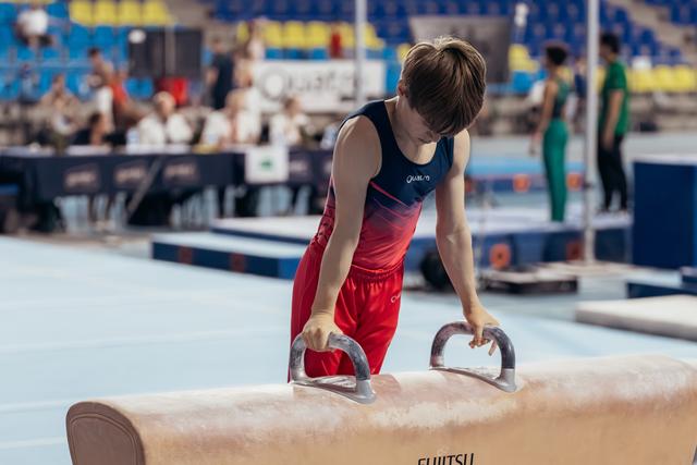 Young male gymnast grips pommel horse handles, leaning forward with intense concentration during a gymnastics competition.