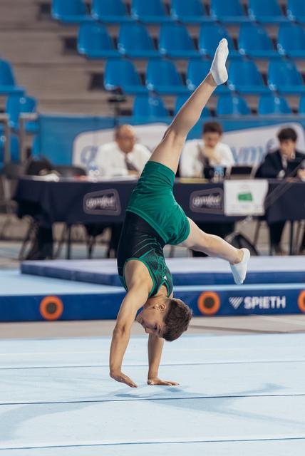 Young male gymnast performs a one-armed handstand with legs split on the floor exercise mat during competition.