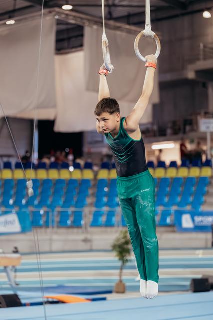 Young male gymnast hangs from still rings with intense focus, wearing green and black leotard in a competition arena.