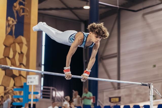 Young male gymnast performs a straddle hold on the horizontal bar, face intense with concentration, in a competition hall.