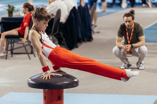 Focused young male gymnast performs a straddle hold on a mushroom while a coach watches attentively in the background.
