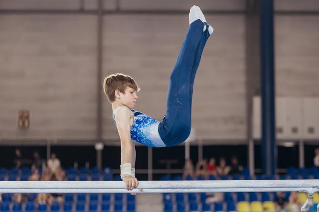 Focused young male gymnast performs an inverted hold on parallel bars, legs raised high, wearing a blue leotard.