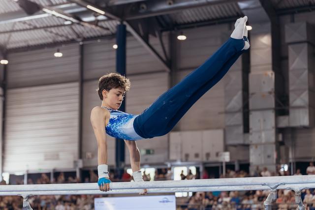 Young male gymnast performs a straddle hold on parallel bars, showing intense focus and strength in a sports hall.