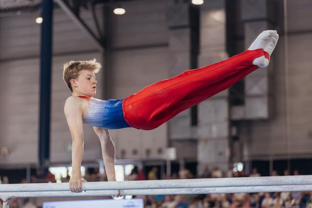 Young male gymnast performs a straddle hold on parallel bars, expression focused and determined, indoor arena.