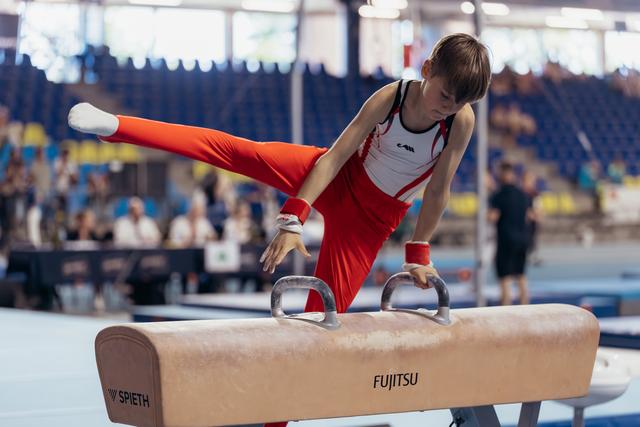 Young male gymnast performs on pommel horse with intense focus, legs extended, wearing red and white uniform.