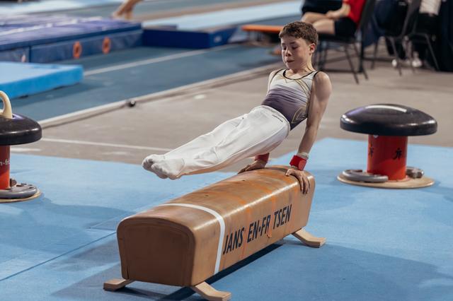 Young male gymnast performs a scissor hold on the pommel horse, concentrated expression, in a blue gymnasium.