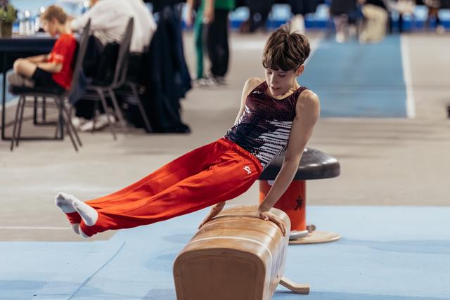Young male gymnast performs a straddle hold on pommel horse, focused expression, at an indoor gymnastics event.