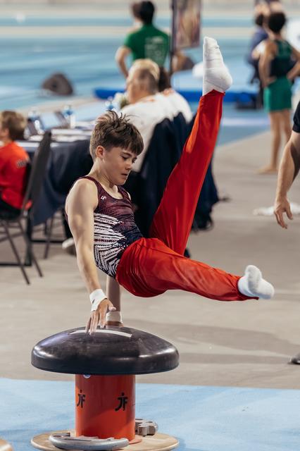 Young male gymnast performs a scissor element on the pommel horse, legs split high, expression calm and concentrated.